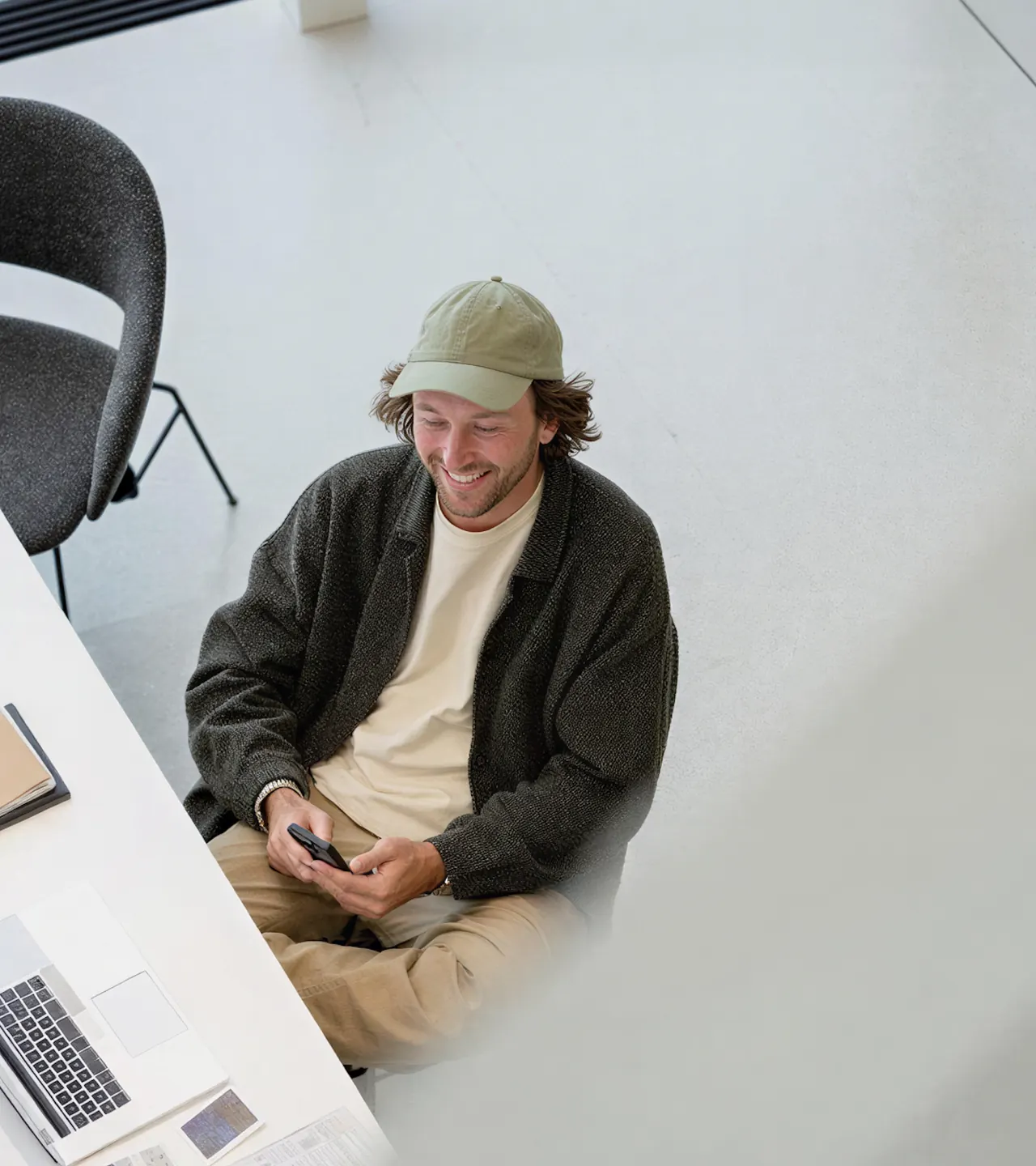 Man at a white desk in a bright studio, looking at a phone beside a laptop and notebook